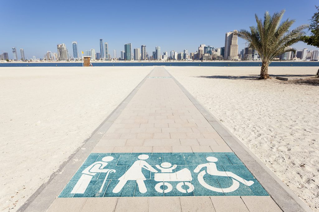 image of bike trail in the middle of sandy desert leading to a city. The trail is marked with a person using a cane, a person pushing a child in a stroller, and a person using a wheelchair.