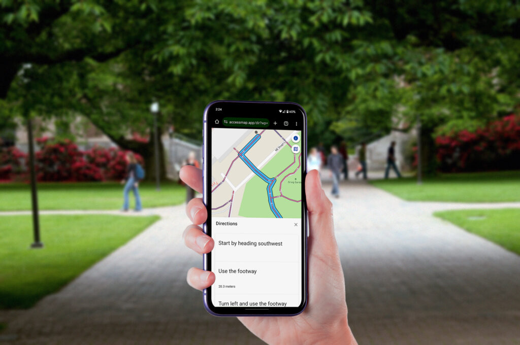 Image of a hand holding a phone with a pathway and trees in the background. The phone screen shows AccessMap Multimodal step by step instructions.
